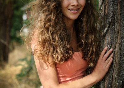 Girl with curly brown hair leaning on a tree in nature.